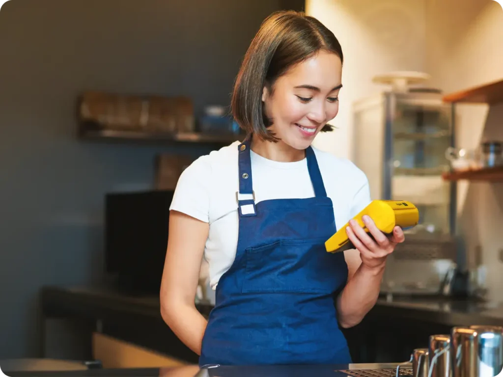 Mulher jovem de cabelo curto usando avental azul em restaurante segurando a maquininha Yelly Pro, ilustrando os planos e taxas da Yelly